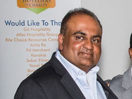 A man in a suit stands near a “Hoteliers for Charity” banner listing various sponsors and supporters, with text partially visible.