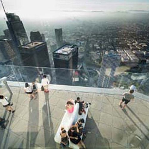 People are on an elevated observation deck with a glass railing, overlooking a city skyline under clear skies.