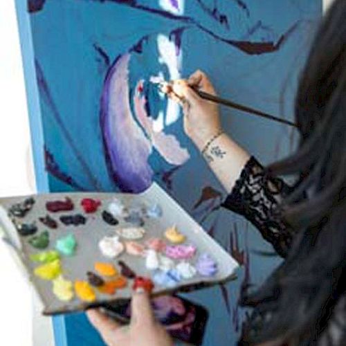 A person is painting on a canvas using a brush, holding a palette with various paint colors. They appear to be creating an abstract artwork.