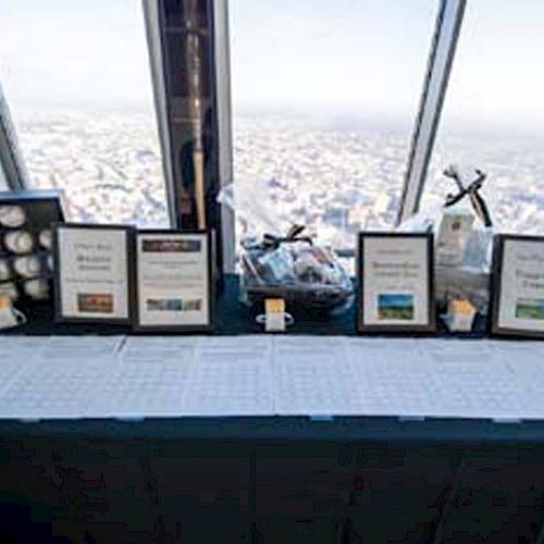 A table with a black cloth has framed signs, golf balls, and a gift basket displayed by a window overlooking a cityscape, ending the sentence.