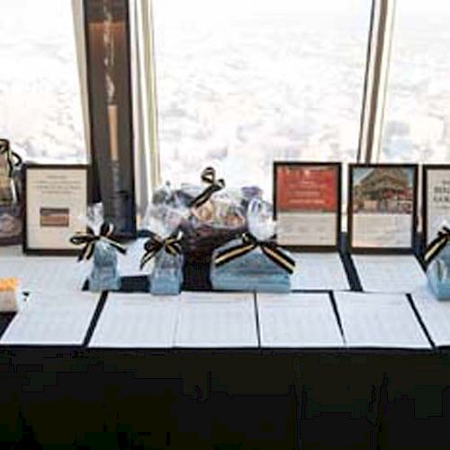 A table displays framed pictures, papers, and gift bags tied with ribbons near a window with a panoramic city view ending the sentence.