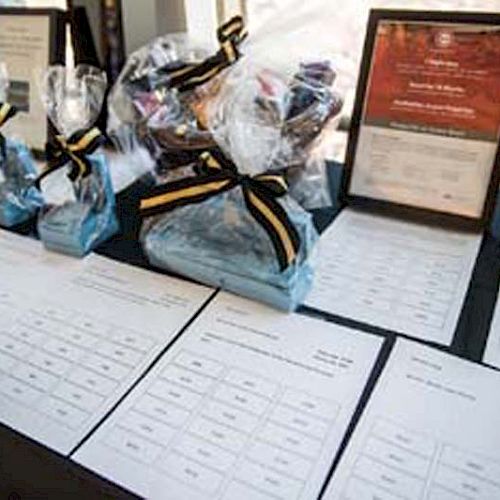 The image shows a table with gift bags, tied with ribbons, and documents, including framed certificates and sheets with printed lists ending the sentence.