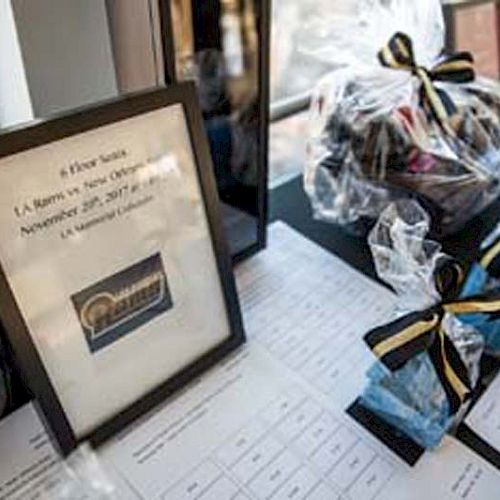 The image shows a framed sign, gift bags with ribbons, and a few documents on a table, seemingly for an event or auction.