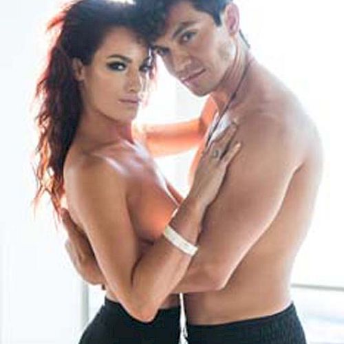 A topless man and woman stand close together, facing the camera, with a bright background of windows behind them.