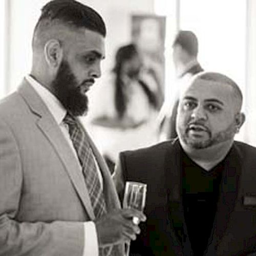 Two men are engaged in a conversation at what appears to be a formal event, both holding glasses, with other attendees in the background.