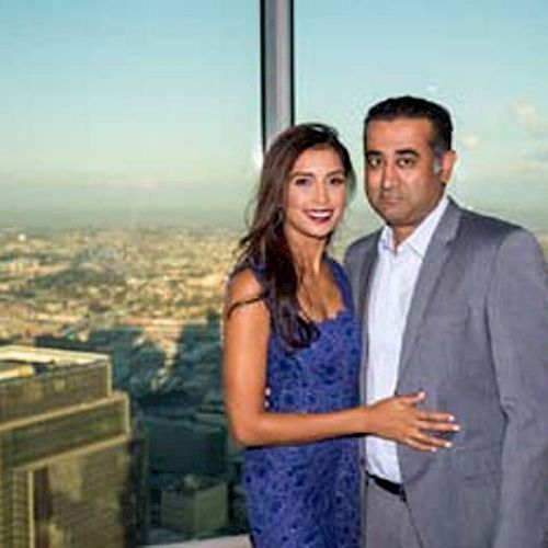 A couple is posing in front of a large window with a city skyline view in the background. The woman is wearing a blue dress, while the man wears a gray suit.