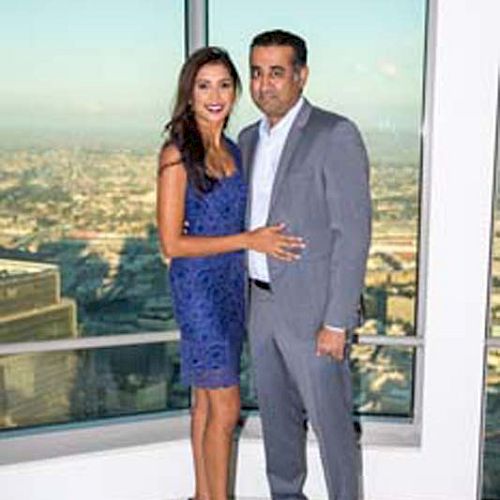 A couple dressed elegantly stands in front of a large window with a cityscape in the background.