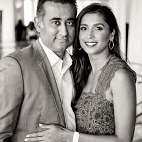 A black and white photo of a smiling couple; the man is in a suit, and the woman is in a lace dress. They appear to be in a formal setting.