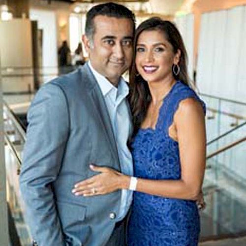 A well-dressed couple poses for a photo in an indoor setting with modern decor, with the woman wearing a blue dress and the man in a grey suit.