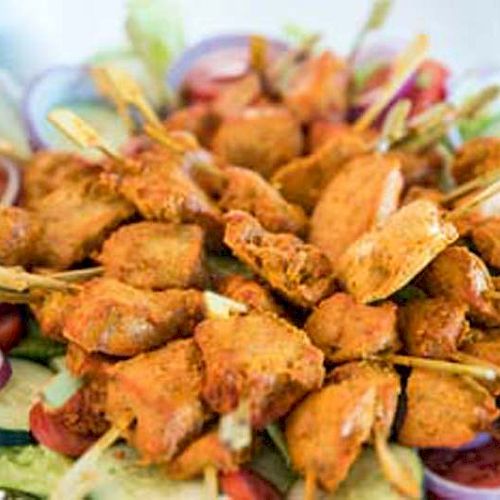 A platter with skewers of marinated grilled meat, possibly chicken, surrounded by sliced cucumbers, tomatoes, and red onion rings on a bed of lettuce.