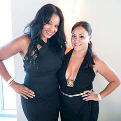 Two women in black outfits stand side by side, smiling at the camera. They're behind a table with papers and markers.