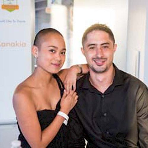 The image shows two people, a woman with a shaved head and a man with short hair, smiling and posing together in a bright indoor setting.