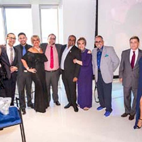 A group of people, dressed formally, standing together inside a modern room with large windows. The group has smiles, mingles, and poses for the photo.