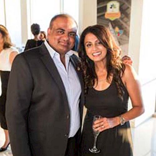 A man and woman are posing for a photo at an event, both dressed formally. The woman is holding a glass of wine and they appear to be smiling.