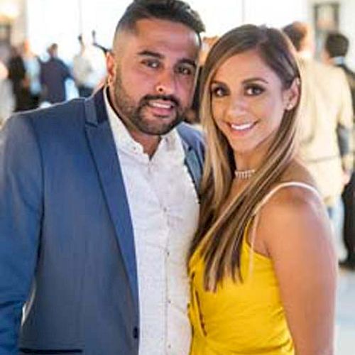 A man in a suit and a woman in a yellow dress are posing together in a well-lit, indoor setting surrounded by several people.