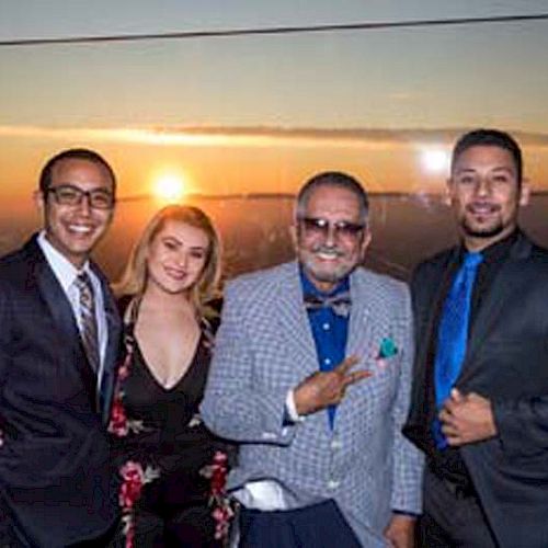 Four people in formal attire pose in front of a sunset backdrop; one flashes a peace sign, while the others smile.