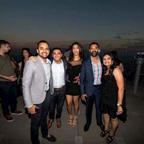 A group of people are posing for a photo at an outdoor evening event with a scenic view in the background.