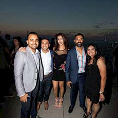 Five people are posing for a photo at what looks like an outdoor event during the evening. They are dressed in formal or semi-formal attire.