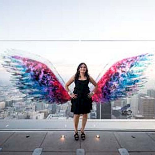 A person stands in front of a glass wall with colorful painted wings, giving the appearance of having wings, with a cityscape in the background.