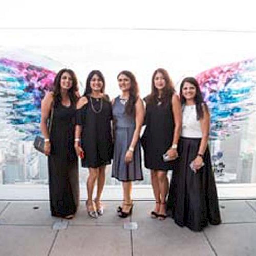 Five women are standing side by side in front of a colorful wing mural, with a cityscape visible in the background.