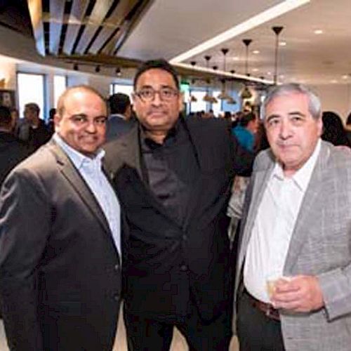 Three men in suits are posing together at an indoor event with other people in the background.