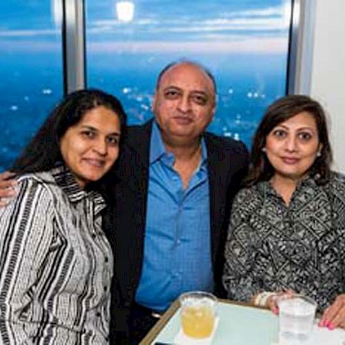 Three people are posing together indoors by a window; one man and two women, smiling with drinks on the table in front of them.