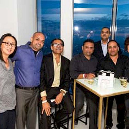 A group of people are posing for a photo at an indoor event with drinks on a table and a view of the evening sky outside.