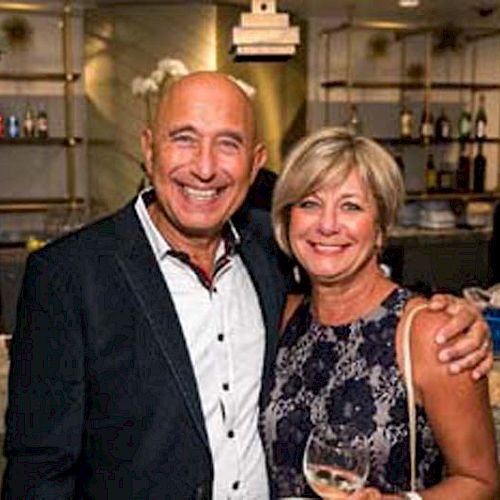 A happy couple stands at a bar, smiling for the camera, the man in a suit and the woman in a lace dress, both holding drinks.