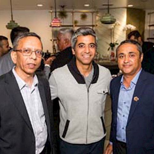 Three men stand together smiling at an indoor gathering with more people in the background.