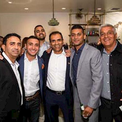 A group of six men is posing together in a well-lit indoor setting, dressed in formal and semi-formal attire, smiling at the camera.