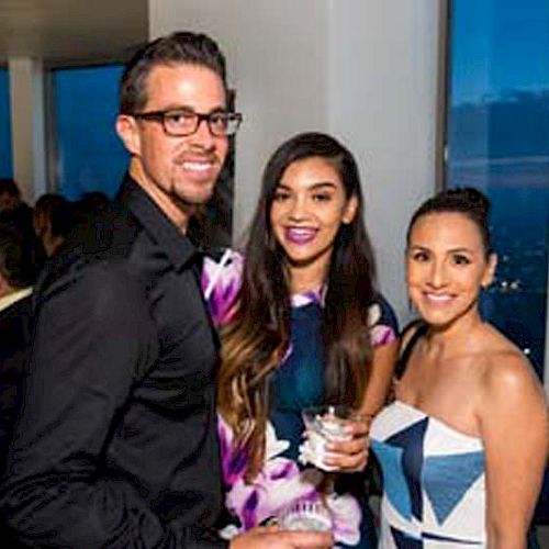 Three people are posing for a photo at an indoor event, smiling and holding drinks, with a city view through large windows in the background.