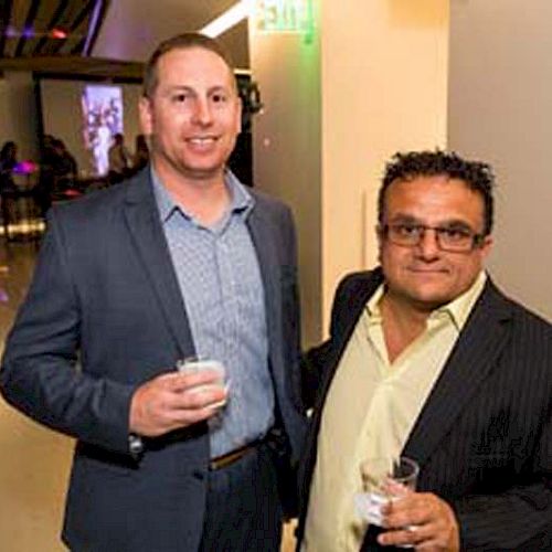 Two men are standing together at an event, holding drinks and smiling; a sign behind them mentions a children's hospital and sponsors.