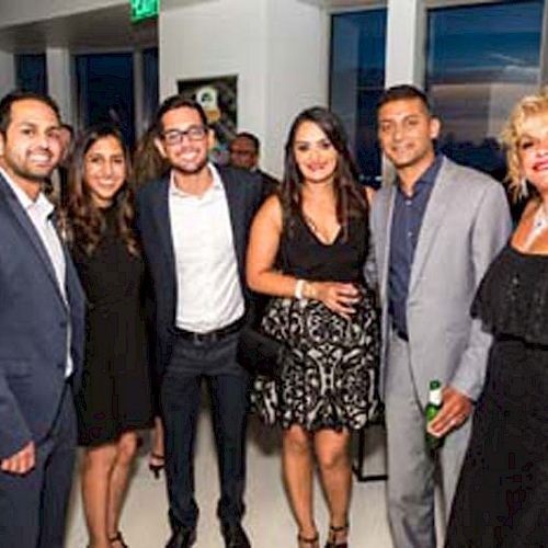 A group of people are posing and smiling at a social event, dressed in formal attire, against a backdrop of large windows and a sunset view.