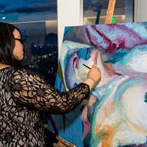 A woman is painting on a canvas at an easel in a well-lit room with large windows showing a nighttime cityscape outside.