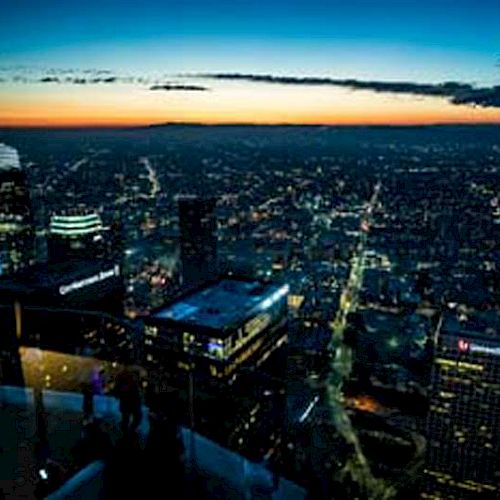 The image shows a cityscape at dusk with illuminated buildings, a darkening sky, and a horizon with an orange-yellow glow from the setting sun.