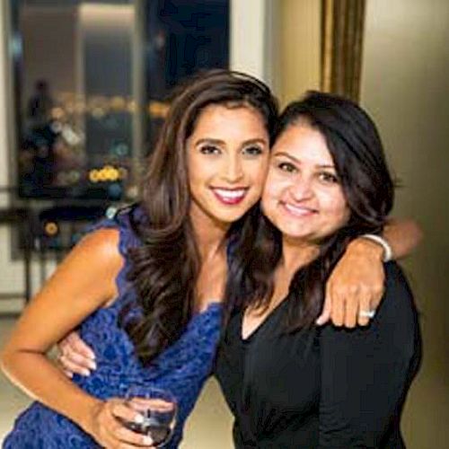 Two women are smiling at the camera in what appears to be an indoor setting. One woman is holding a drink.