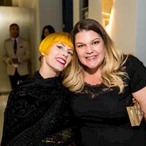 Two women are smiling and posing for a photo at an event; one has short yellow hair, and the other has long blonde hair, both dressed in black.