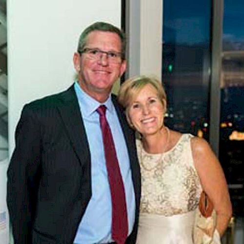 Two people are smiling and posing together at an indoor event. They are formally dressed, with a city view in the background.