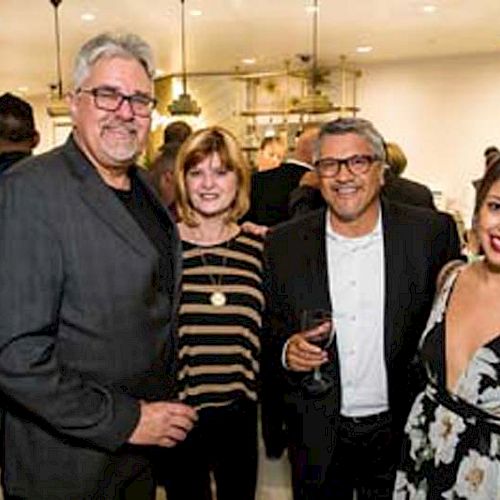 Four people are posing together at a social event, holding drinks and smiling at the camera in a well-lit area.