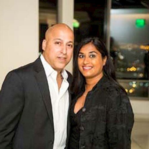 A man and woman are posing together indoors, dressed in formal attire, with a cityscape visible through the windows in the background.