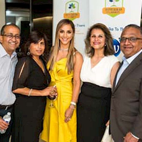 A group of five people posing indoors, with women wearing dresses, one in a yellow dress. They're in front of a banner featuring event sponsors.