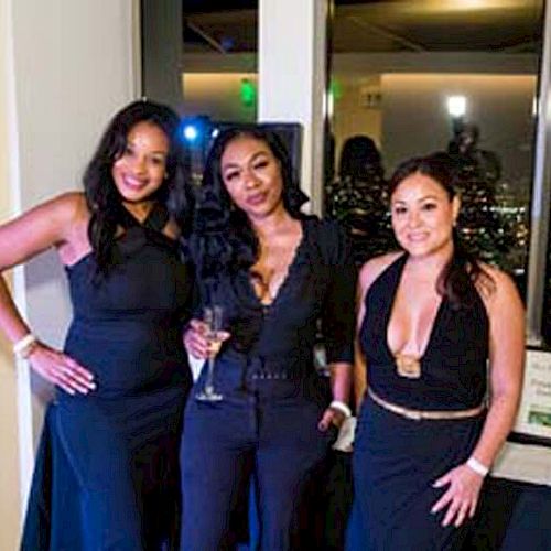 Three women are posing together in formal attire, with city lights visible through the windows behind them.