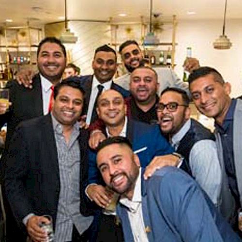 A group of men in formal attire are gathered together, smiling and holding drinks in a lively, well-lit indoor setting.