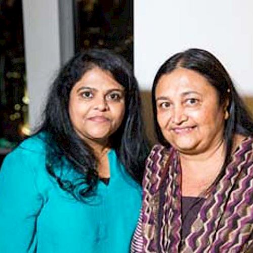 Two women are standing indoors, smiling at the camera. One is wearing a teal top and the other is in a patterned shawl.