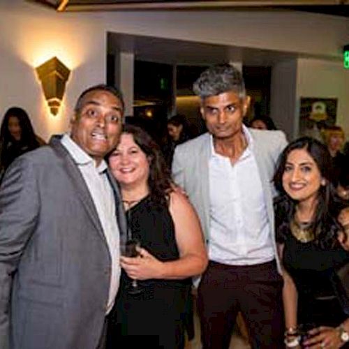 A group of four people dressed in formal attire is posing for a photo at a social event, with other attendees visible in the background.