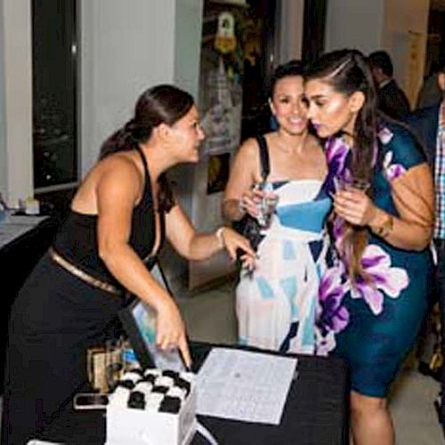 People are gathered at a table at an event. One person is gesturing while others are holding drinks and conversing, appearing to enjoy themselves.