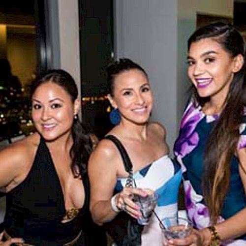 Three women are posing together at a social gathering, holding drinks, with a cityscape visible through the windows behind them.