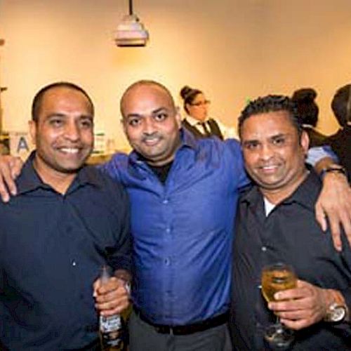 Three men are posing together at a social event, each holding a drink, with a bar and other guests in the background.