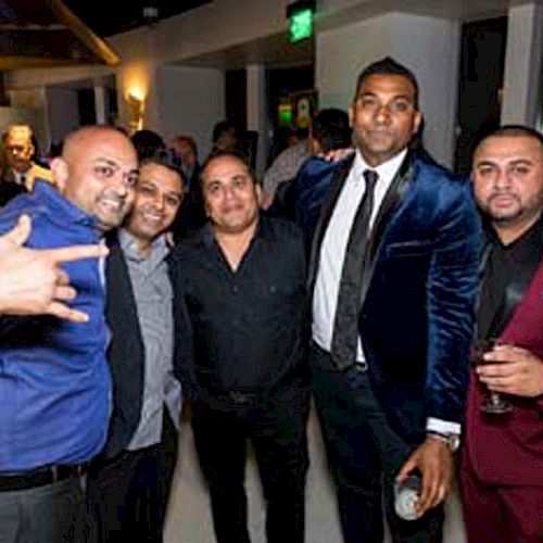 A group of five men dressed in formal attire, posing together at an indoor event with others in the background, smiling and enjoying.