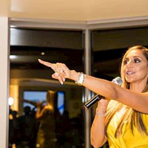 A woman in a yellow dress is holding a microphone and pointing while speaking in a brightly lit room with large windows.